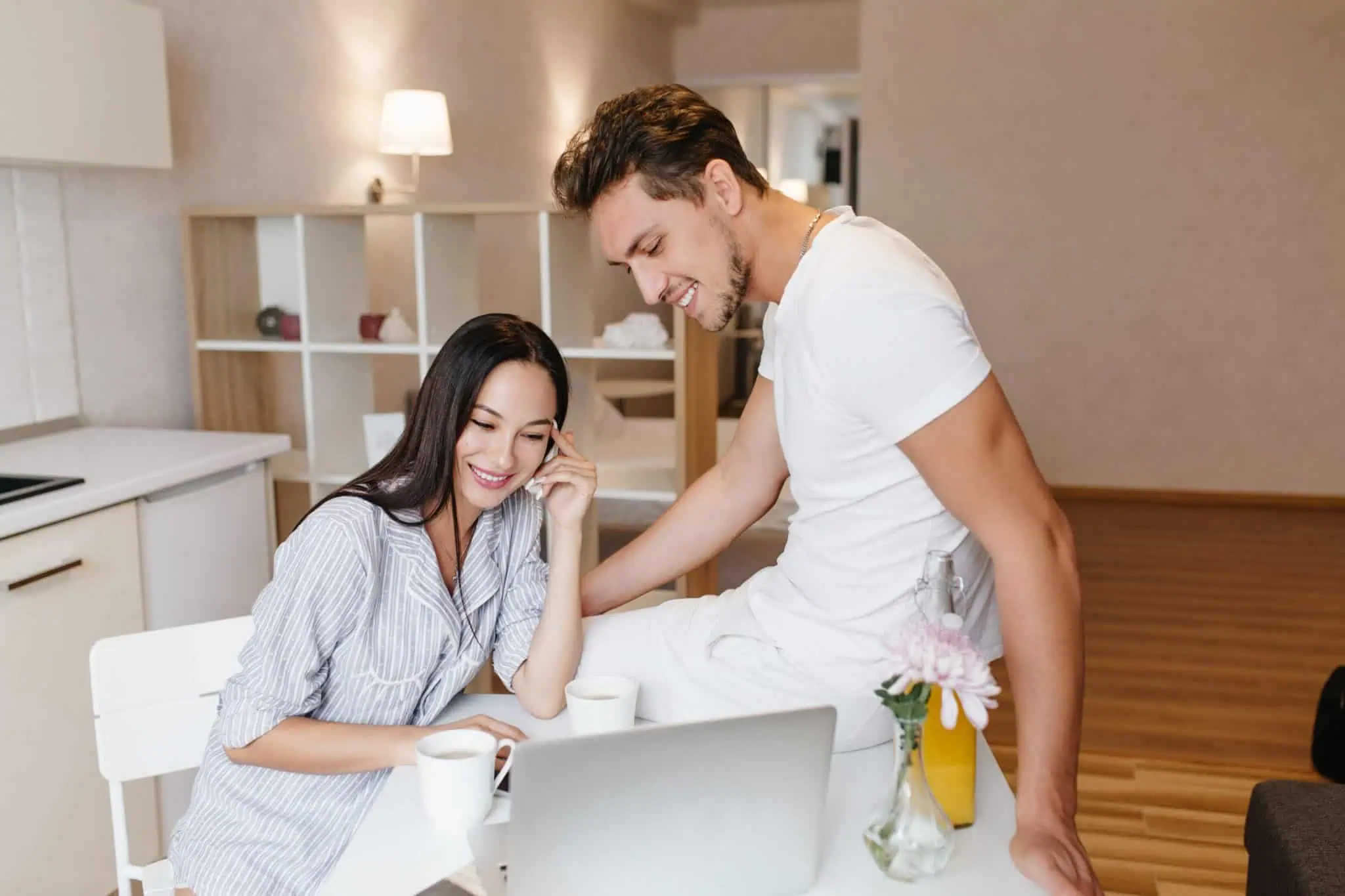 Refined girl with shiny black hair using laptop during breakfast with boyfriend. Indoor portrait of loving couple laughing together and sitting in kitchen with computer.