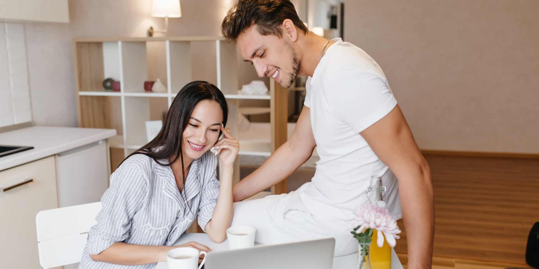 Refined girl with shiny black hair using laptop during breakfast with boyfriend. Indoor portrait of loving couple laughing together and sitting in kitchen with computer.
