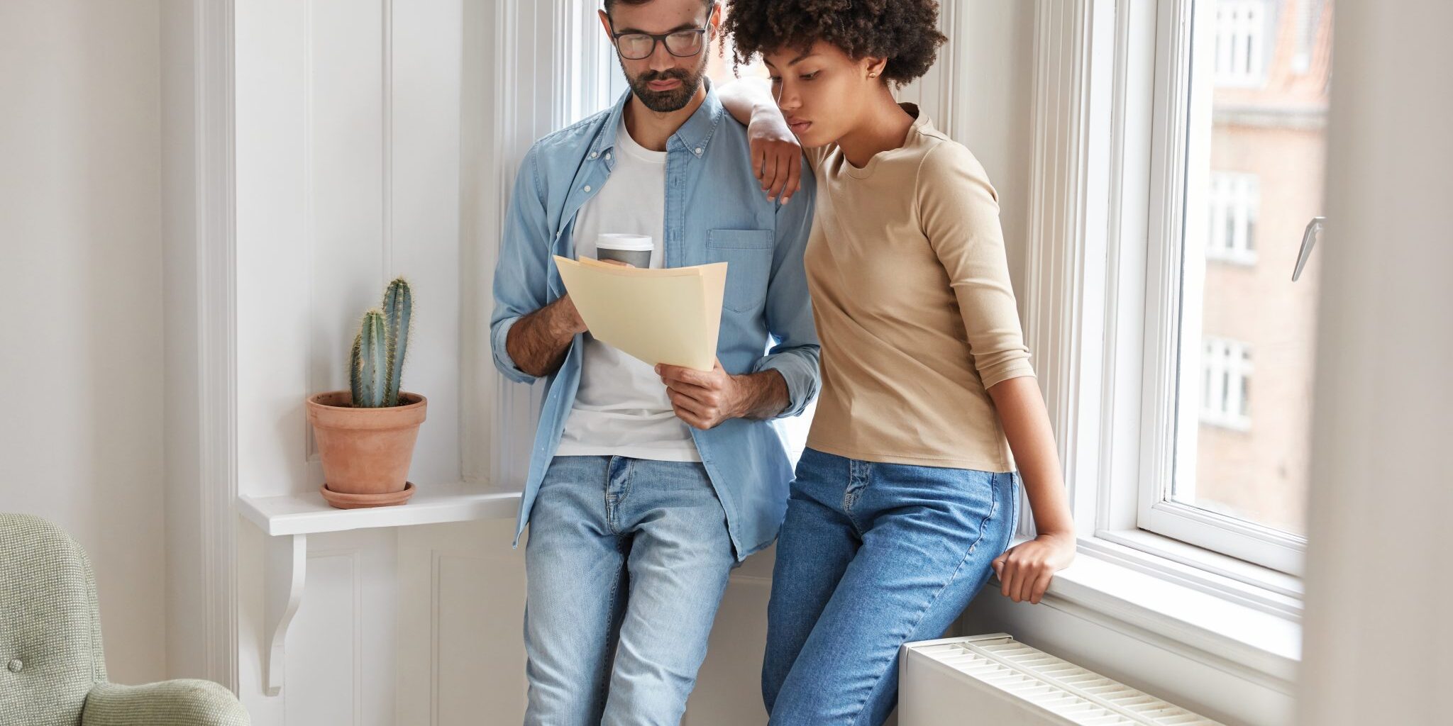 Photo of dark skinned woman and her husband think how satisfy debt, look at bills, secure good bargain with business partner, pose near big window indoor. Entrepreneur want to recieve high profits